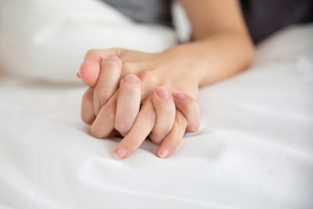 Sex under mensen – er det trygt, og hva bør du vite? Nærbilde av to hender som holder hverandre på hvitt laken, symbol på intimitet, trygghet og kjærlighet under sex.