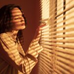 Kvinne i varmt sollys ser ettertenksomt ut gjennom persienner – et øyeblikk av nærvær og egenrefleksjon.