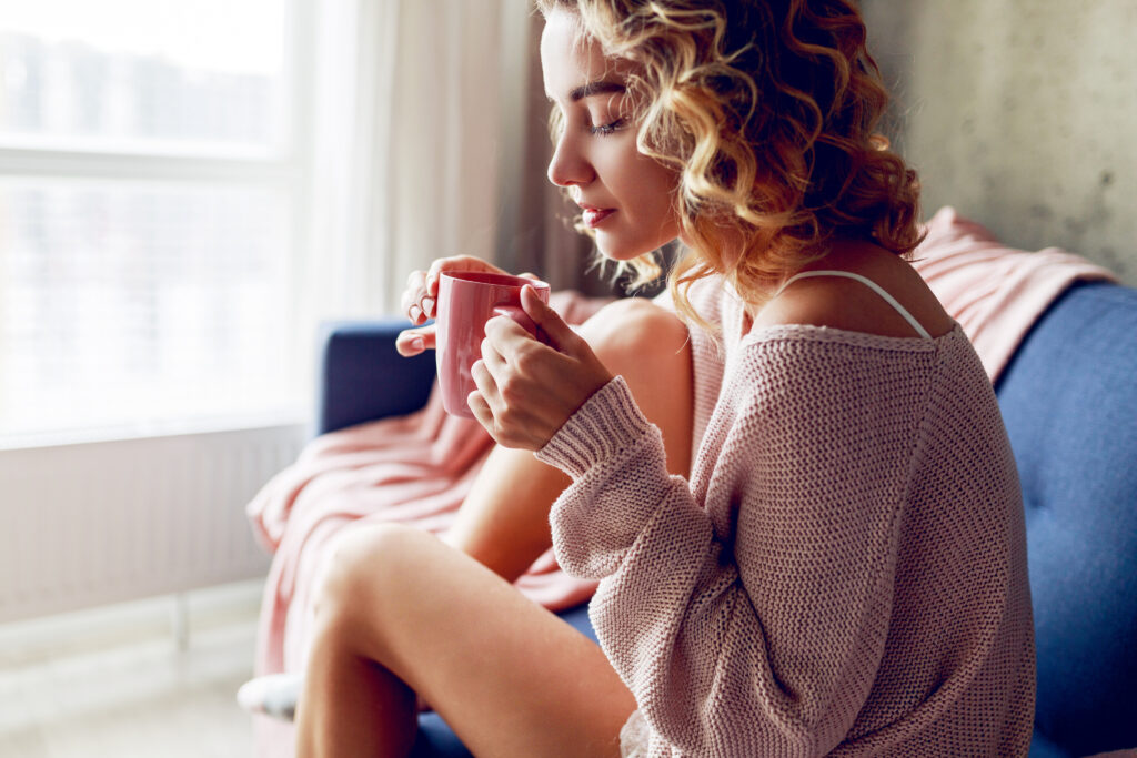 Kvinne i myk rosa genser nyter en stille stund med kaffe, sittende i sofaen med lukkede øyne og et fredelig uttrykk – et øyeblikk av egenomsorg og ro.
